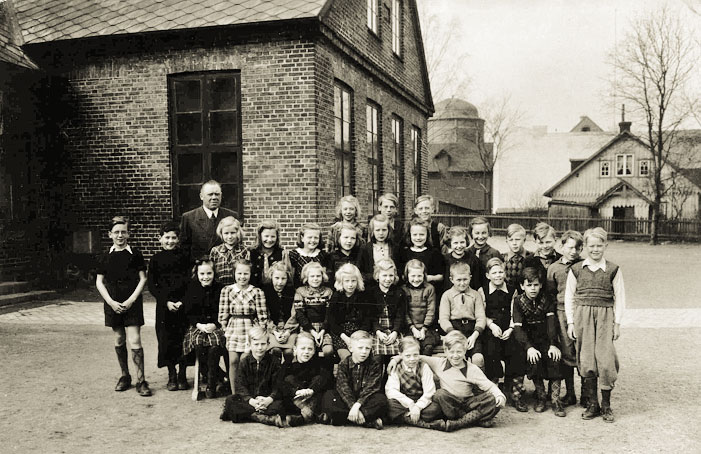 I Esl&ouml;vsbon Susanne Hultmans fotoblogg kan vi l&auml;sa: Den h&auml;r bilden hittade jag bland min pappas gamla foton och den visar den h&ouml;gra gaveln p&aring; &Ouml;stra skolan, men ur en annan vinkel &auml;n p&aring; min bild. L&auml;raren &auml;r f&ouml;rst&aring;s Joel Sallius. L&auml;ngst ner till h&ouml;ger sitter min pappa och skrattar f&ouml;r fullt &aring;t kameran. Foto: Susanne Hultman.