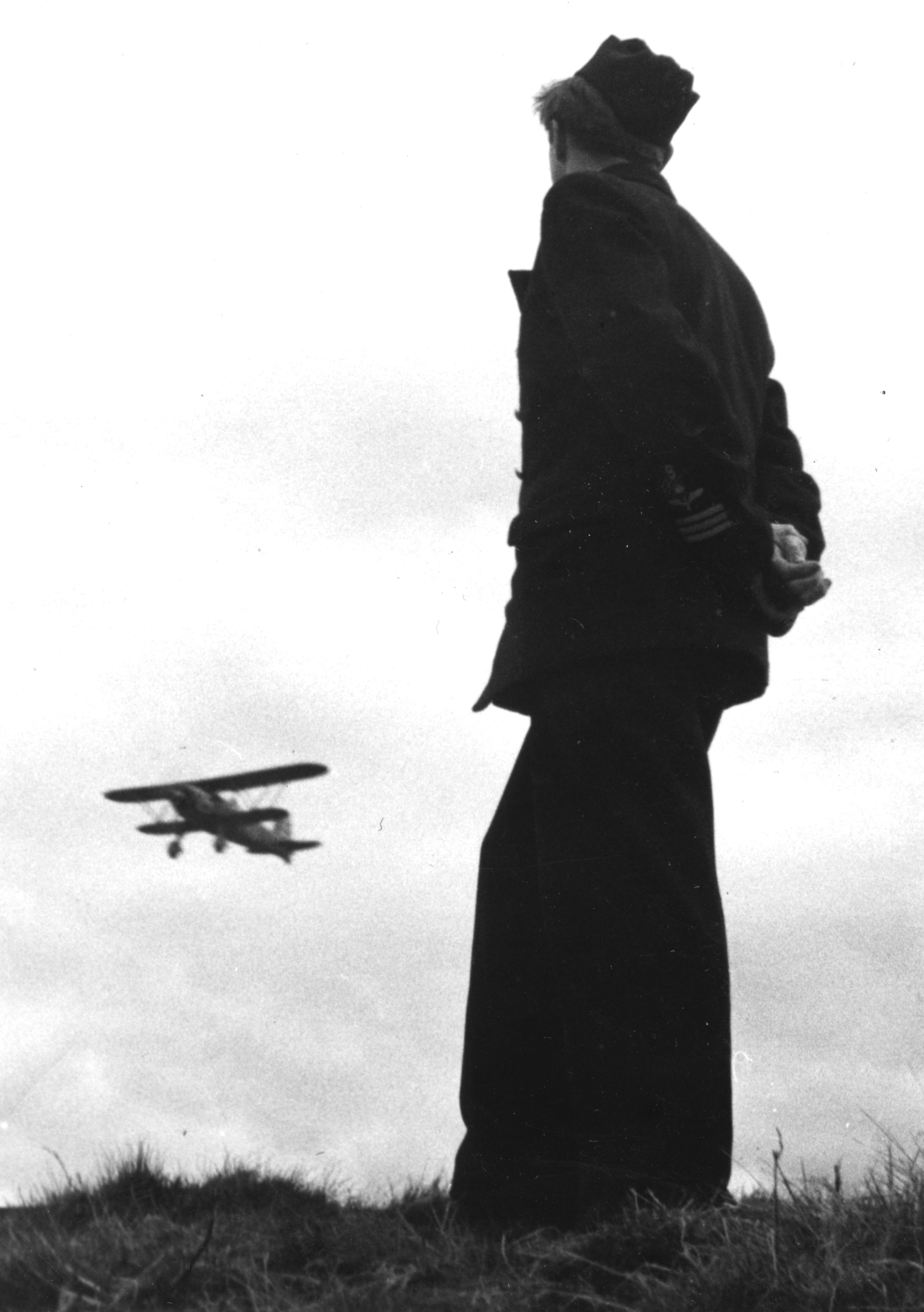 Motiv: En flygsoldat st&aring;rt p&aring; ett flygf&auml;lt betraktar ett milit&auml;rt flygplan i luften, cirka 1943. Ur Kurt Edenstams album 'Minnen fr&aring;n Min kommistid 1940-1944'. Edenholm var stationerad p&aring; Krigsflygskolan F 5 samt F 9. Bild utan bildtext i albumet. Tillh&ouml;r Flygvapenmuseums bildarkiv.